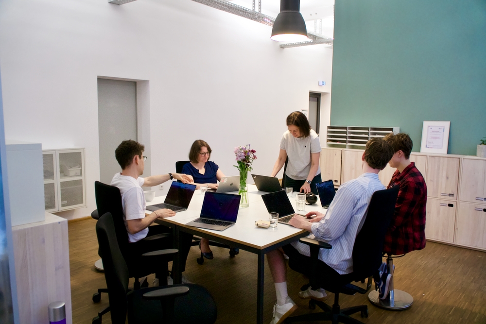 The picture shows five people sitting at a large table with their laptops open in front of them.