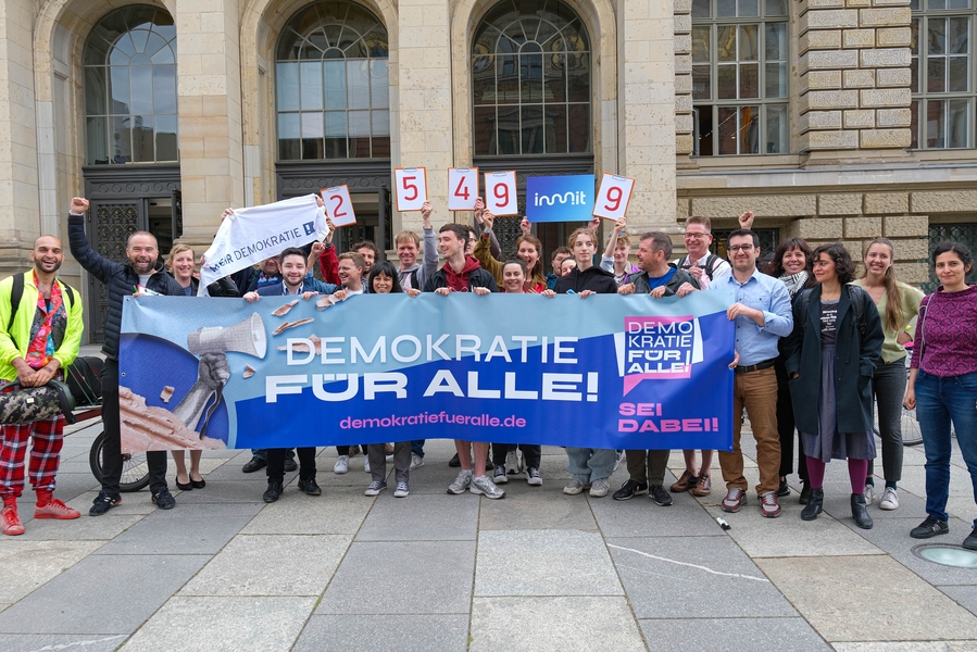 Menschen vor dem Berliner Abgeordnetenhaus halten einen Banner mit der Aufschrift „Demokratie für Alle!“ und Schilder mit der Zahl „25.499“