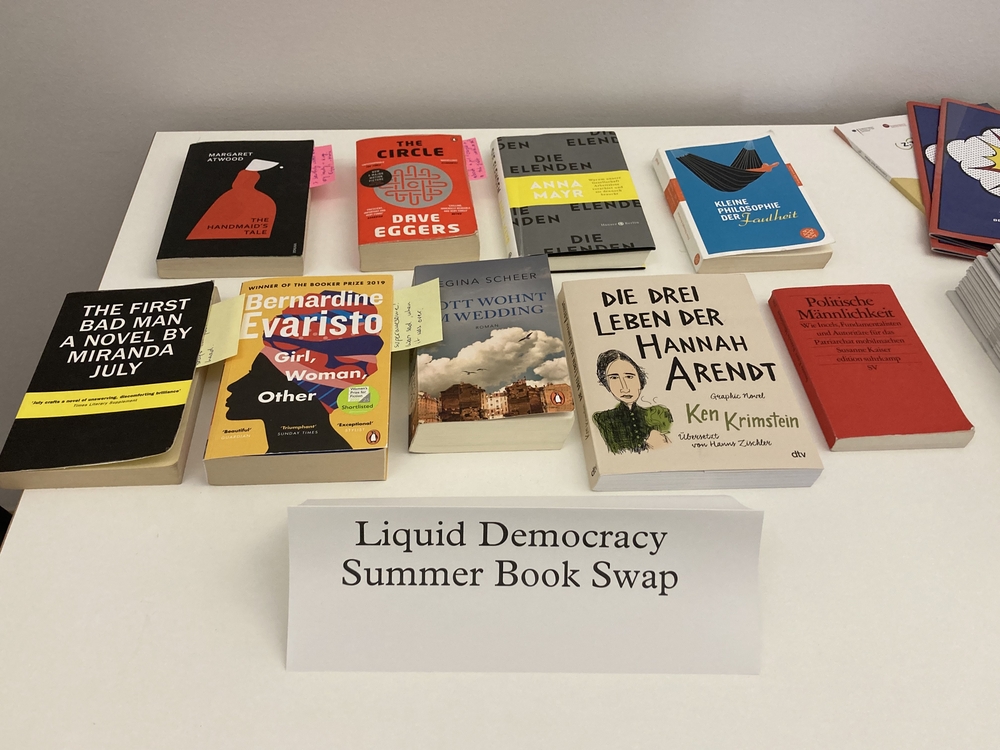 Auf dem Bild sieht man verschiedene Bücher auf einem Tisch liegen. Darunter steht auf einem kleinen Schild: "Liquid Democracy Summer Bookswap"