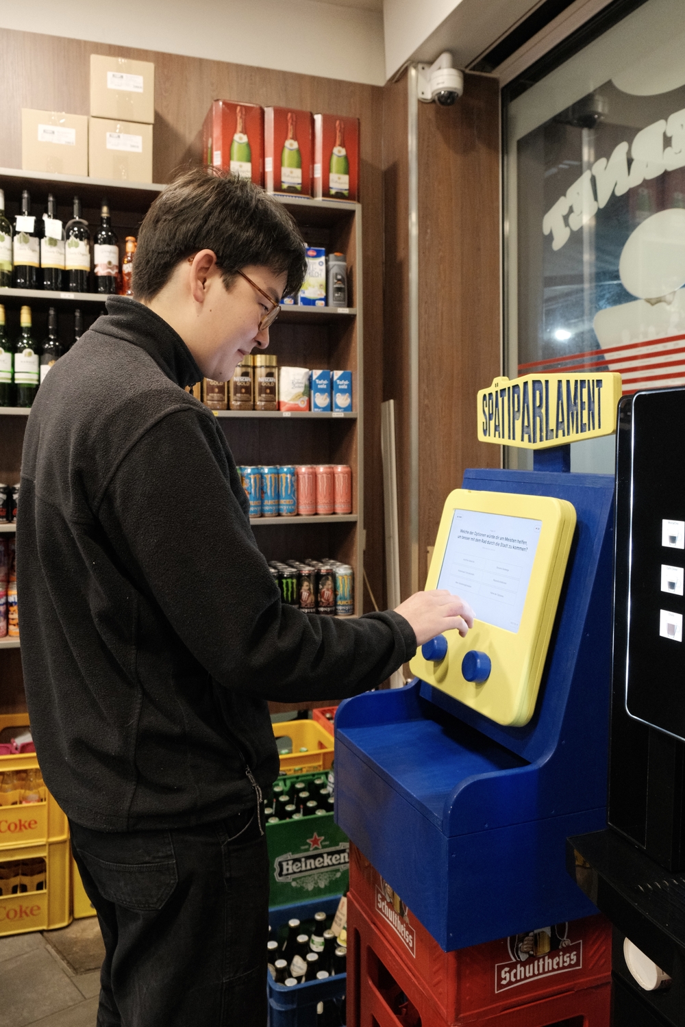 Eine Person bedient ein interaktives Terminal mit der Aufschrift „Spätiparlament“ in einem Berliner Späti. Das Gerät mit Touchscreen steht auf Getränkekisten zwischen Regalen mit Wein, Getränkedosen und anderen Produkten, während die Person eine Eingabe a
