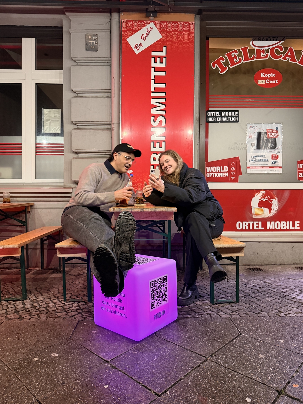 Zwei Personen sitzen nachts an einem Holztisch vor einem Kiosk in Berlin. Beide schauen gemeinsam auf das Display. Im Vordergrund leuchtet ein auffälliger, violetter Würfel mit einem großen QR-Code und deutschem Text.