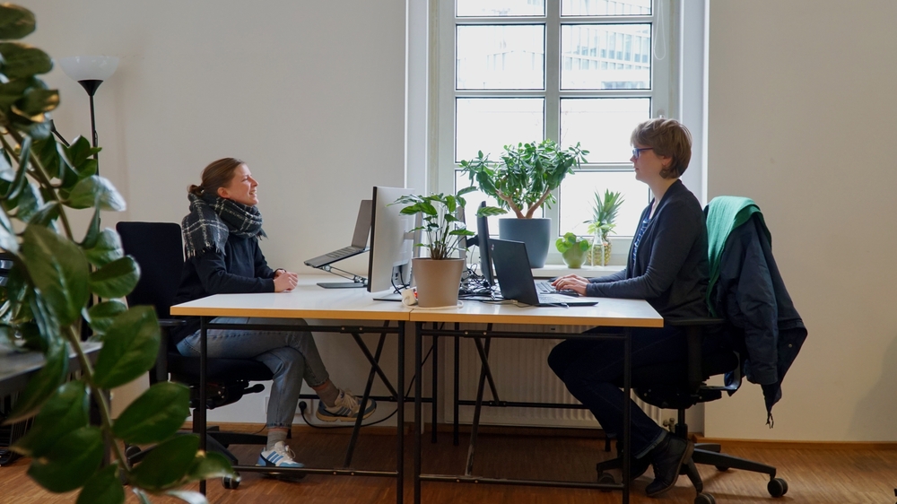 Auf dem Bild sind Rine und Katharina jeweils vor ihren Laptops an einem großen weißen Tisch im Büro zu sehen. Im linken unteren Eck sieht man ein paar Pflanzenblätter, die verschwommen sind.