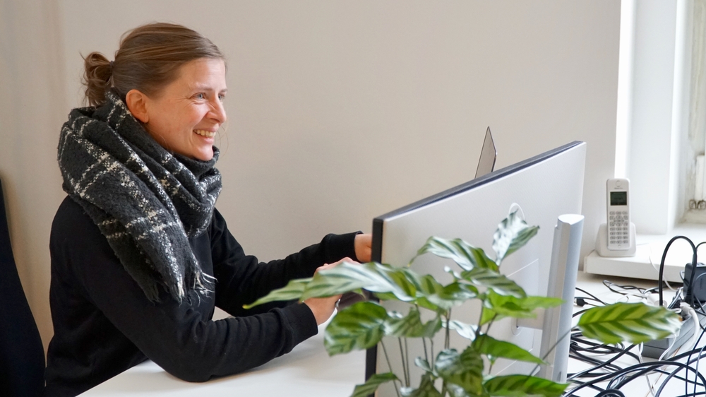 Auf dem Bild ist Rine lächelnd an ihrem Schreibtisch zu sehen. Vor ihr steht ein großer Monitor und eine Pflanze.