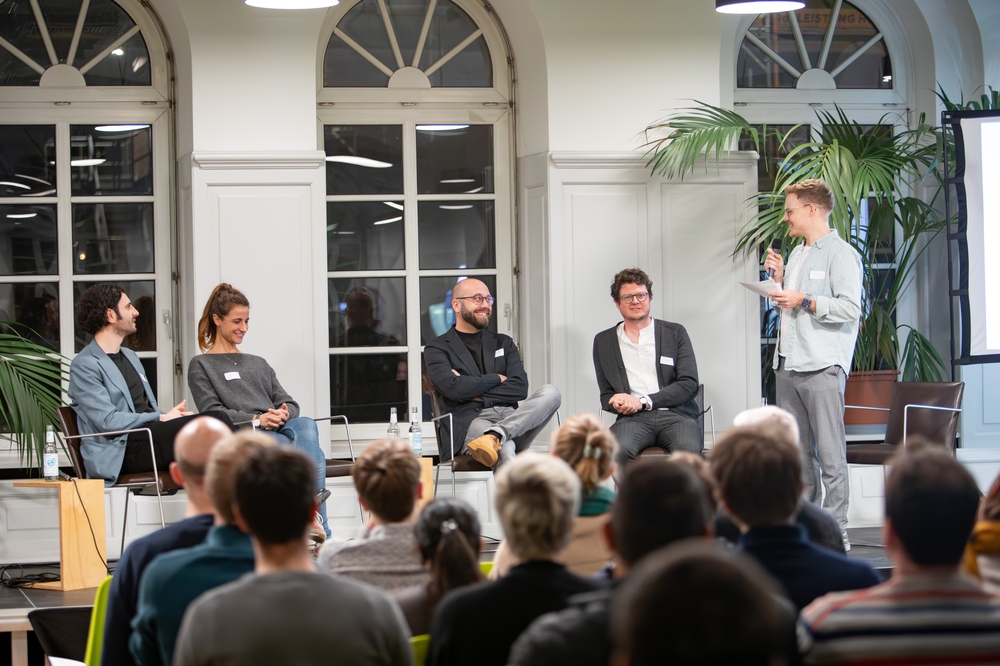 Auf dem Bild ist ein Panel zu sehen. Vier sprecher:innen sitzen auf stühlen und haben einen amüsierten Gesichtsausdruck. Daneben steht ein Moderator mit Mikrofon in der Hand.