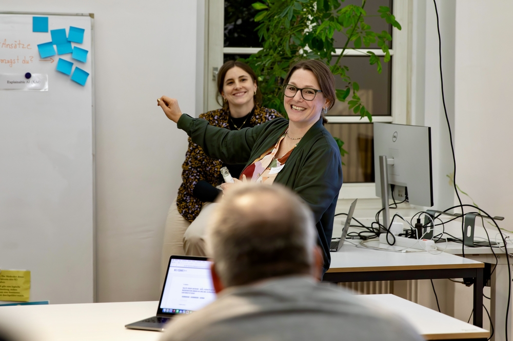 Das Bild zeigt eine lächelnde Person im Büro. Sie hebt ihre Hand und zeigt auf eine Tafel mit blauen Klebeblättern. Im Hintergrund sind Tische, Bildschirme und eine Zuschauerin.
