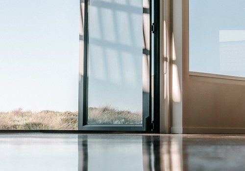Auf der Fotografie ist eine offene Glastür abgebildet, die von einem leeren Raum mit sich spiegeldem Boden nach draußen führt. Draußen ist eine trockene wieße und blauer Himmel zu sehen.