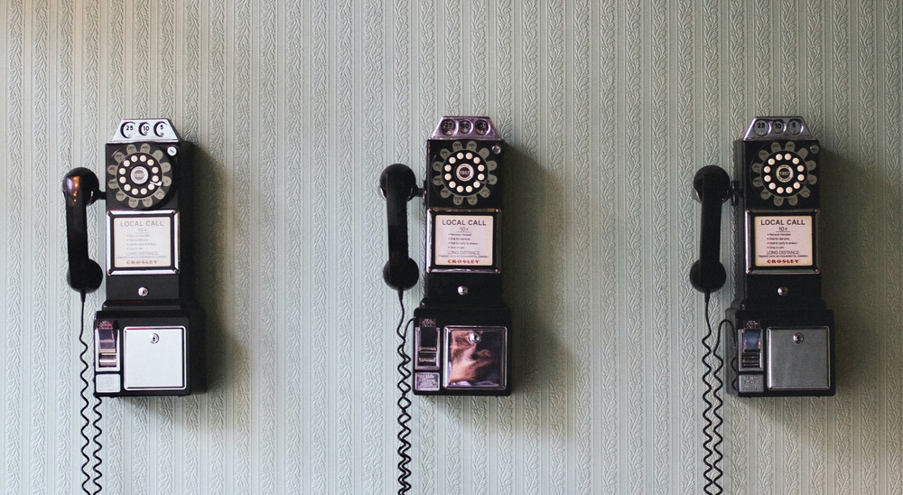 Auf der Fotografie sind 3 alte Telefone abgebildet, die an einer tapezierten weißen Wand hängen.