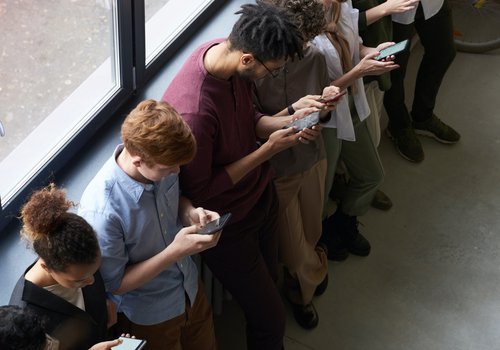 Auf dem Bild ist eine Schlange von Menschen abgebildet, die mit dem Rücken zu einem Fenster stehen und alle in ihre Smartphones schauen.