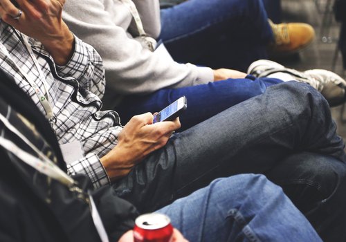 Auf dem Bild sind die Oberkörper und Beine von mehreren sitzenden Personen zu sehen. Die Person in der Mitte des Ausschnitts schaut auf ihr Smartphone.