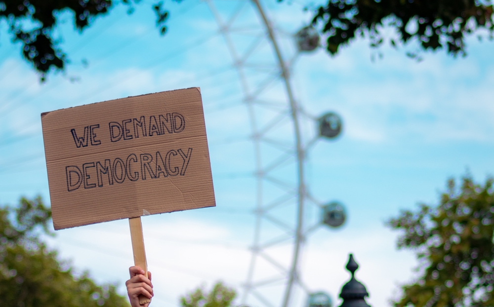 Auf dem Bild ist eine Hand zu sehen, die ein Schild mit der Aufschrift "We demand Democracy" in die Höhe hällt. Im Hintergrund ist verschwommen ein Rieserad und blauer Himmel zu sehen.