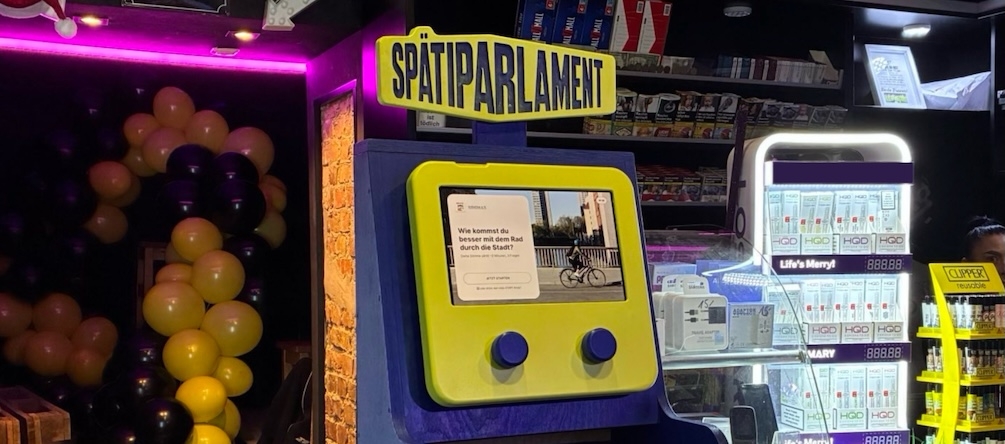 Gelb-blaues interaktives Display mit der Aufschrift 'SPÄTIPARLAMENT' und einem Bildschirm, der eine Frage zum Radfahren in der Stadt zeigt, umgeben von Luftballons und einem beleuchteten Verkaufsregal mit Produkten.