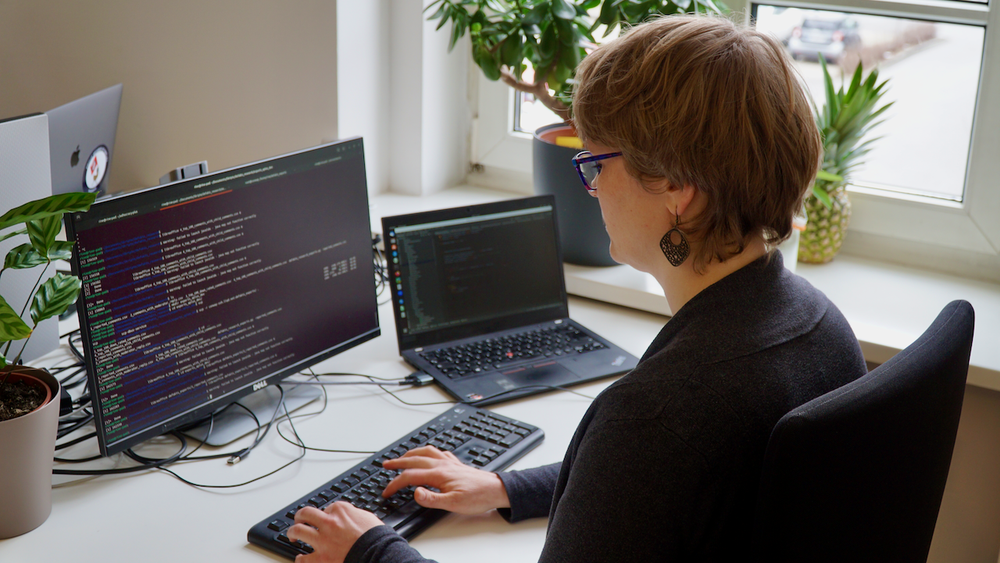 Auf dem Bild ist Katharina von schräg hinten an ihrem Schreibtisch zu sehen. Auf dem Bildschirm ihres Monitors und Laptops ist Code abgebildet. Sie tippt etwas in die Tastatur.